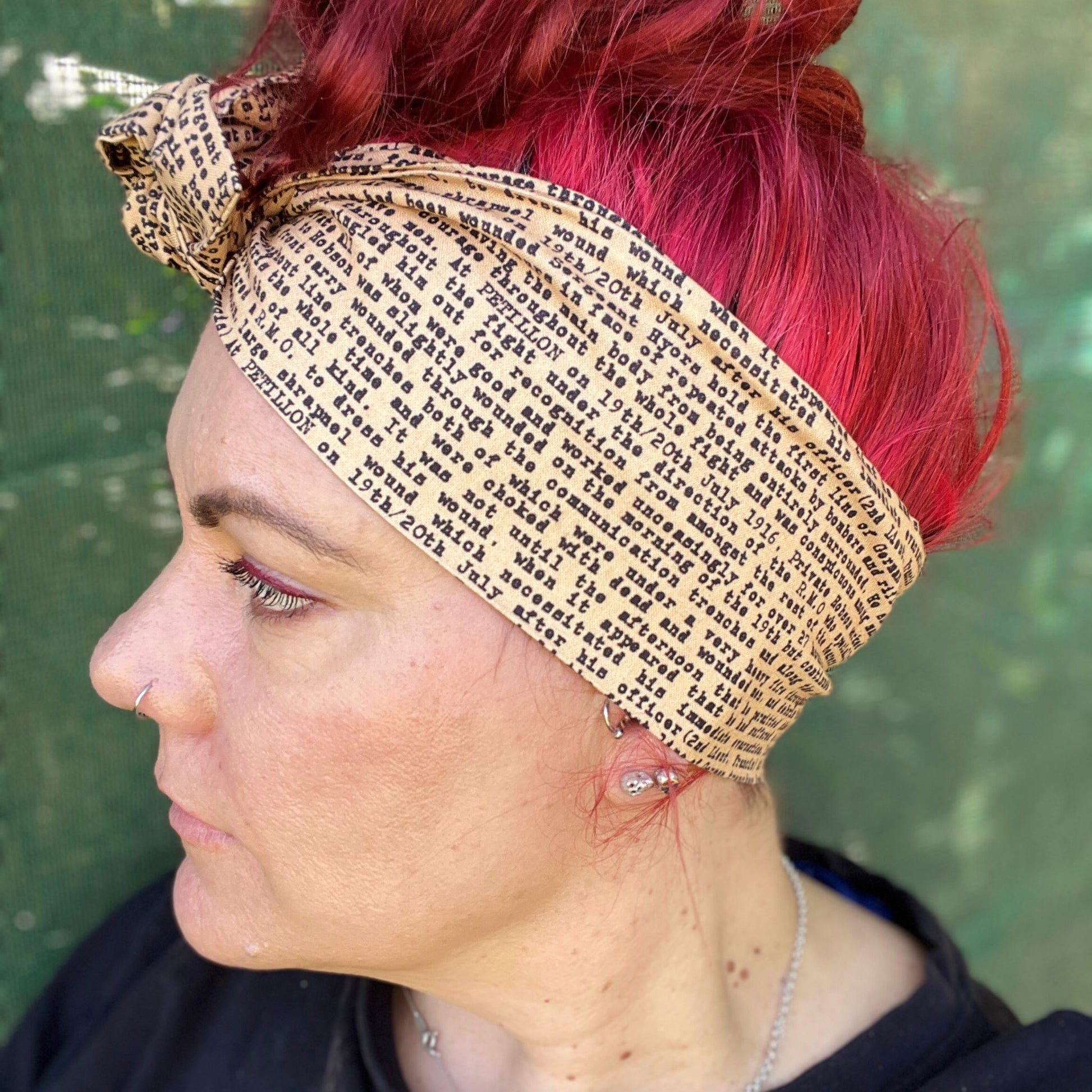 In this image, a woman sports a vibrant red hairstyle, complementing it with a wire headwrap that features an ANZAC soldier's letter a unique print. Adding a historical touch to the headwrap’s design. The headwrap is tied at the top, giving it a bow-like appearance, which accentuates its stylish and laid-back vibe. The woman is looking down, showcasing the headwrap as the standout accessory.