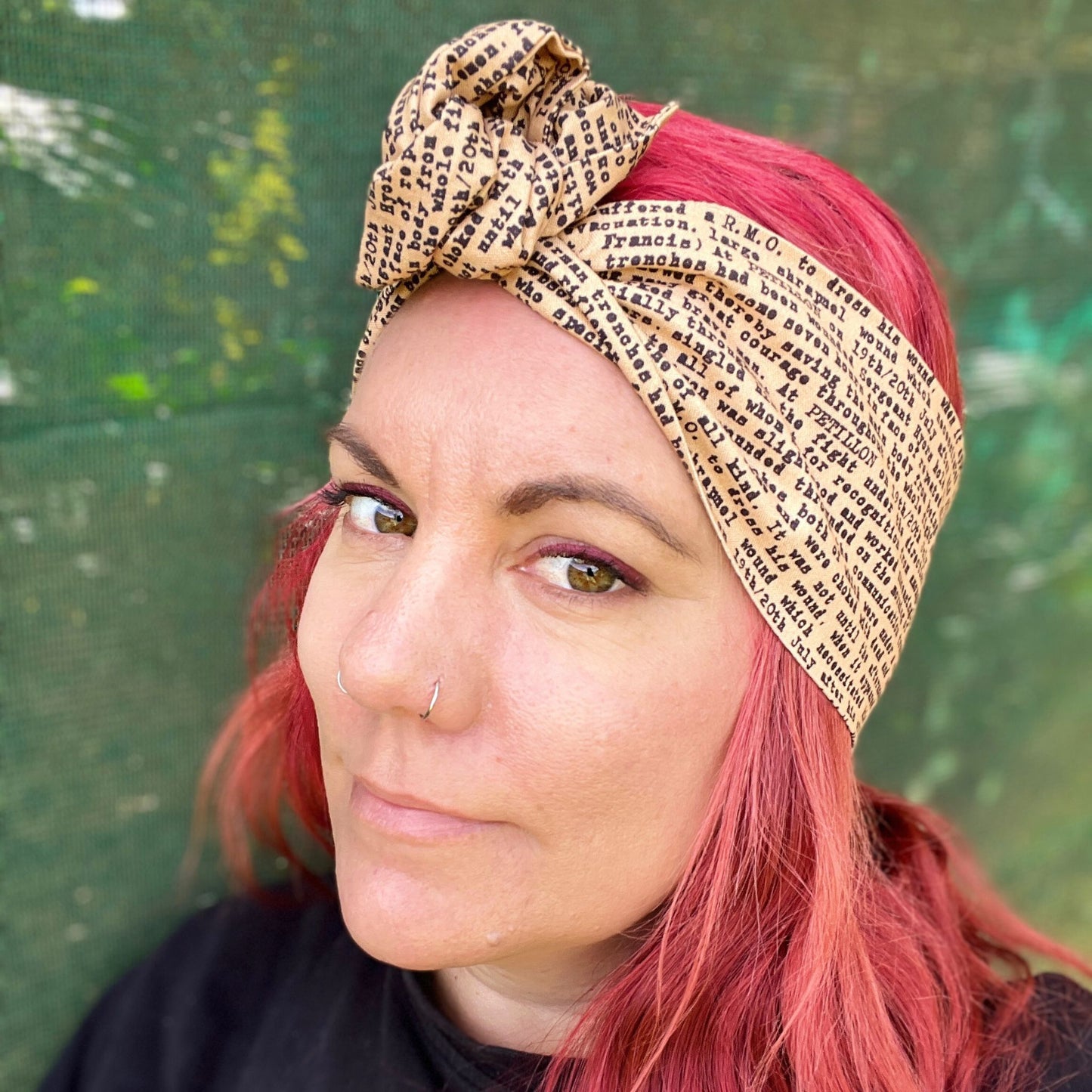 In this image, a woman sports a vibrant red hairstyle, complementing it with a wire headwrap that features an ANZAC soldier's letter a unique print. Adding a historical touch to the headwrap’s design. The headwrap is tied at the top, giving it a bow-like appearance, which accentuates its stylish and laid-back vibe. The woman is looking down, showcasing the headwrap as the standout accessory.