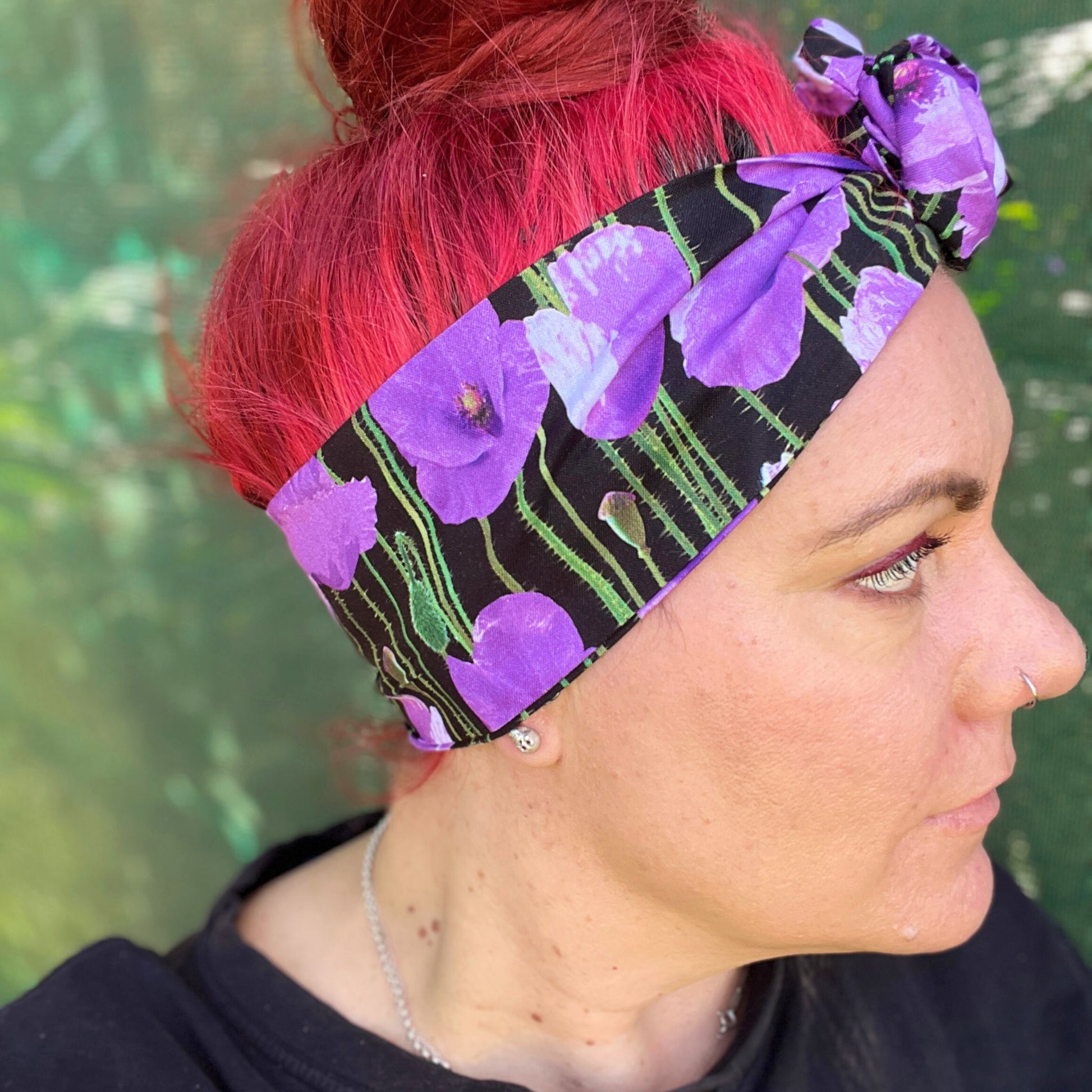 showcasing a wire headwrap adorned with bold purple poppies against a black background. The headwrap is tied in a charming knot atop her head, lending a playful and stylish look. The vivid purple of the flowers stands out