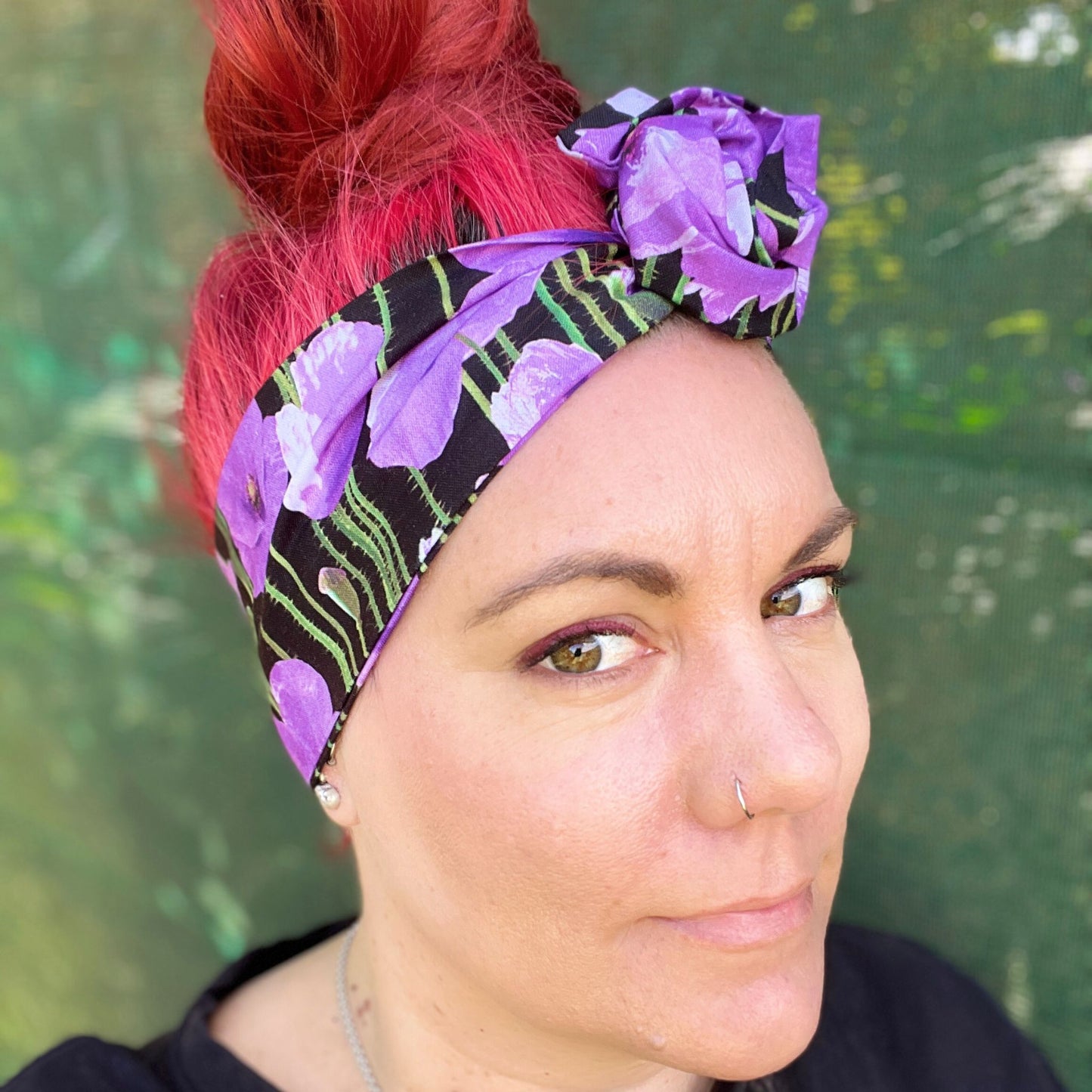 showcasing a wire headwrap adorned with bold purple poppies against a black background. The headwrap is tied in a charming knot atop her head, lending a playful and stylish look. The vivid purple of the flowers stands out