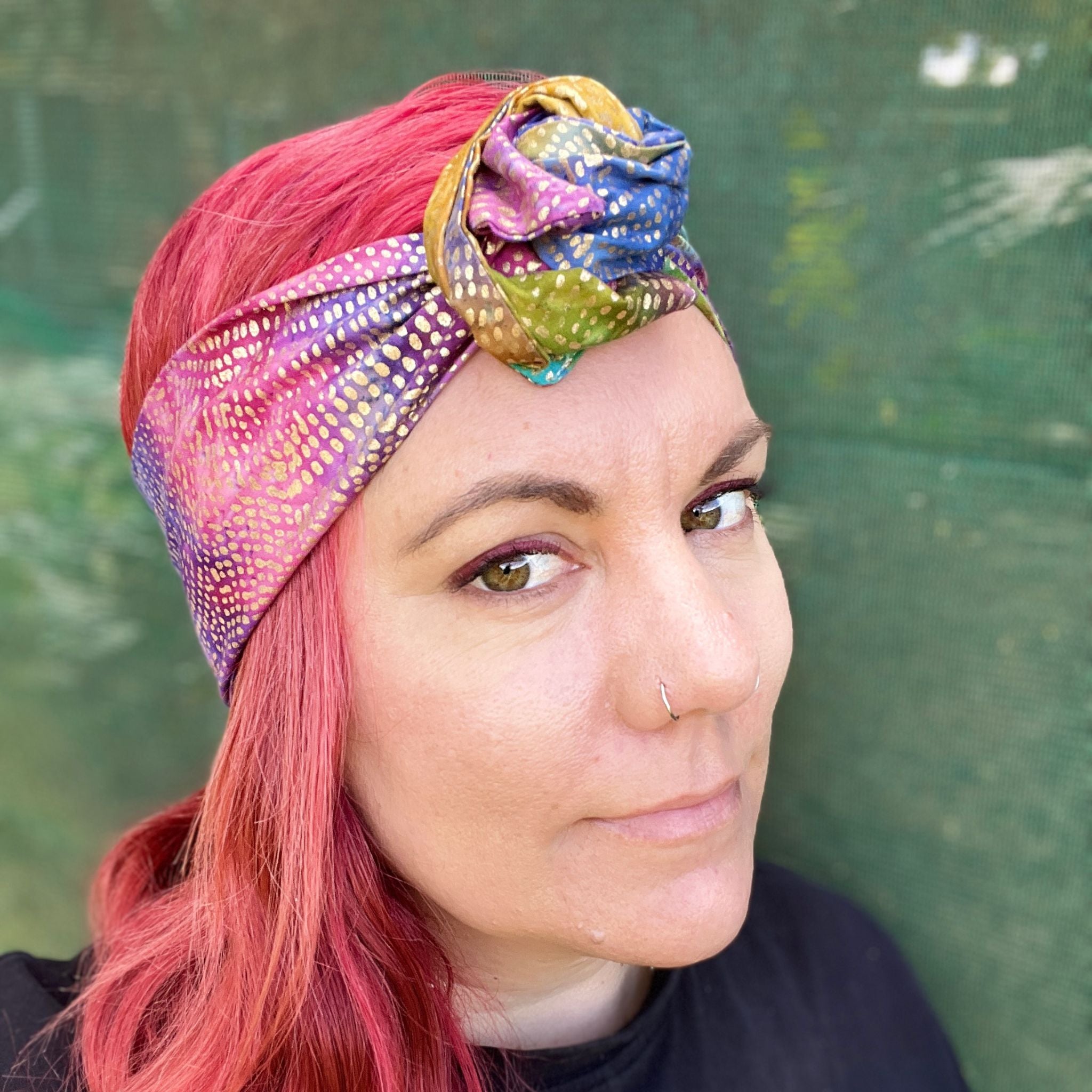 wire headwrap featuring a radiant metallic sheen with a gradient of colors, resembling a rainbow effect. The headwrap is artfully tied in a knot at the side of her head, lending a touch of whimsy to the overall look. 